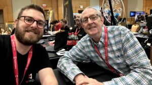 An individual from Magic Media and a conference attendee share a cheerful moment in a selfie at the Pocket Gamer Connects conference 2023, with the busy event atmosphere and gaming stations visible in the background.